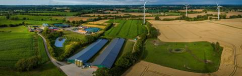 Ilmakuva kesäisestä maaseutumaisemasta. Kuvassa mm. navetta ja tuulivoimaloita.